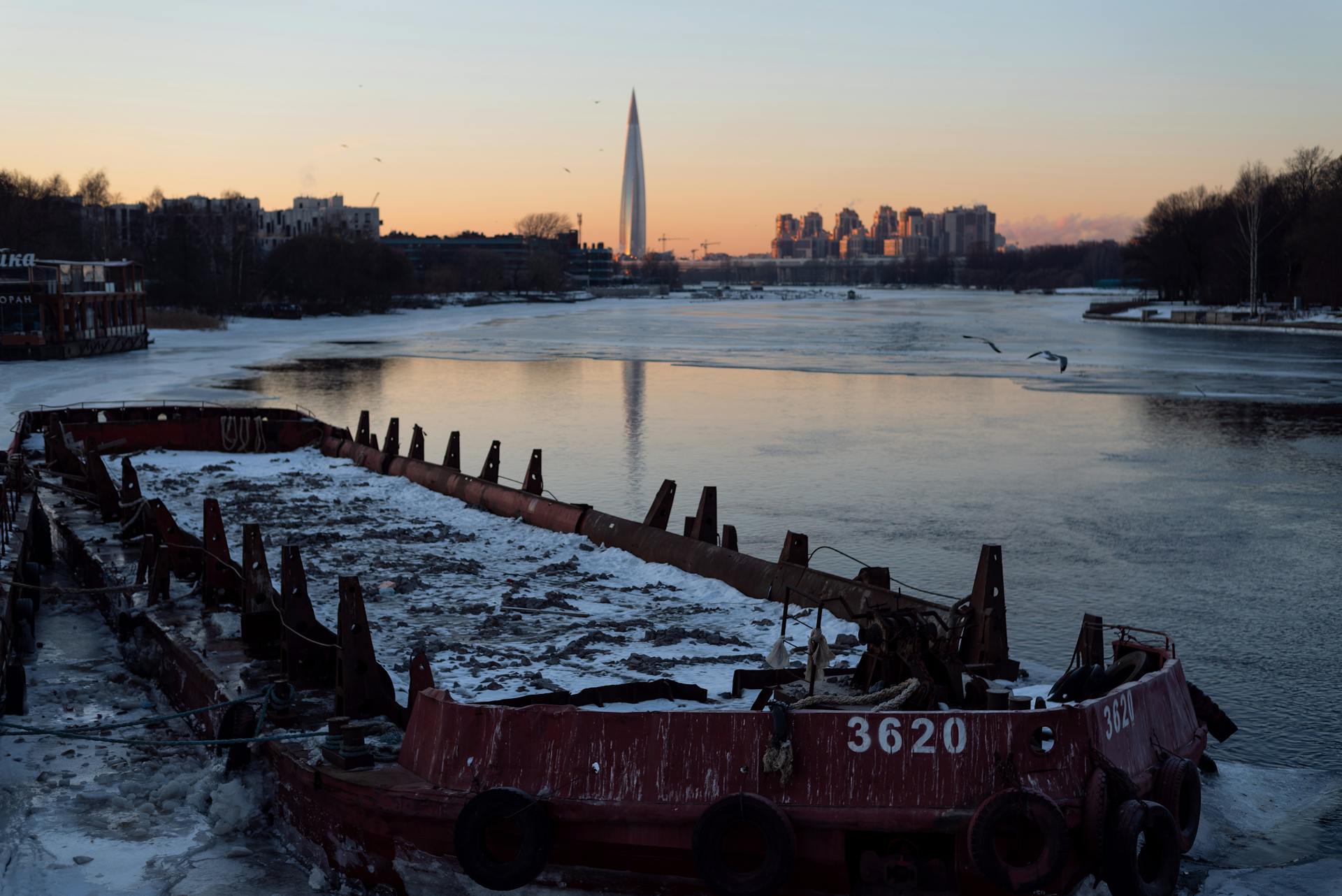 A serene sunset scene featuring a shipwreck on a frozen river with a cityscape and tower in the background.