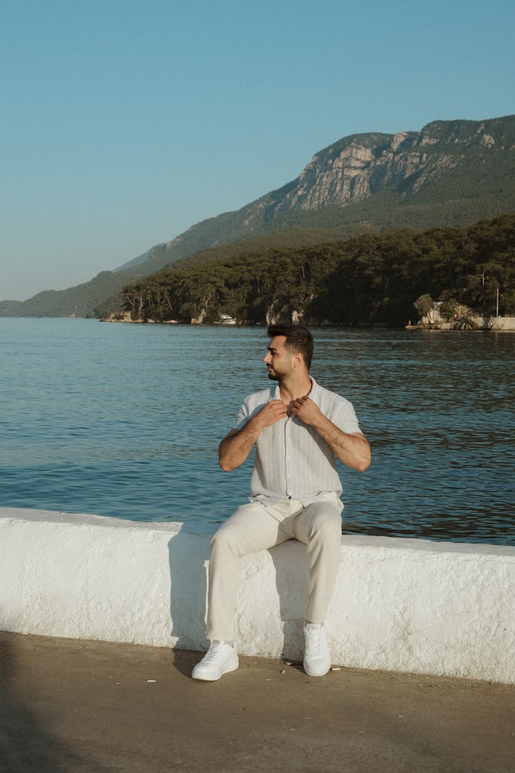 Young Man In White Short Sleeve Shirt And Pants Sitting At A Sea Waterfront