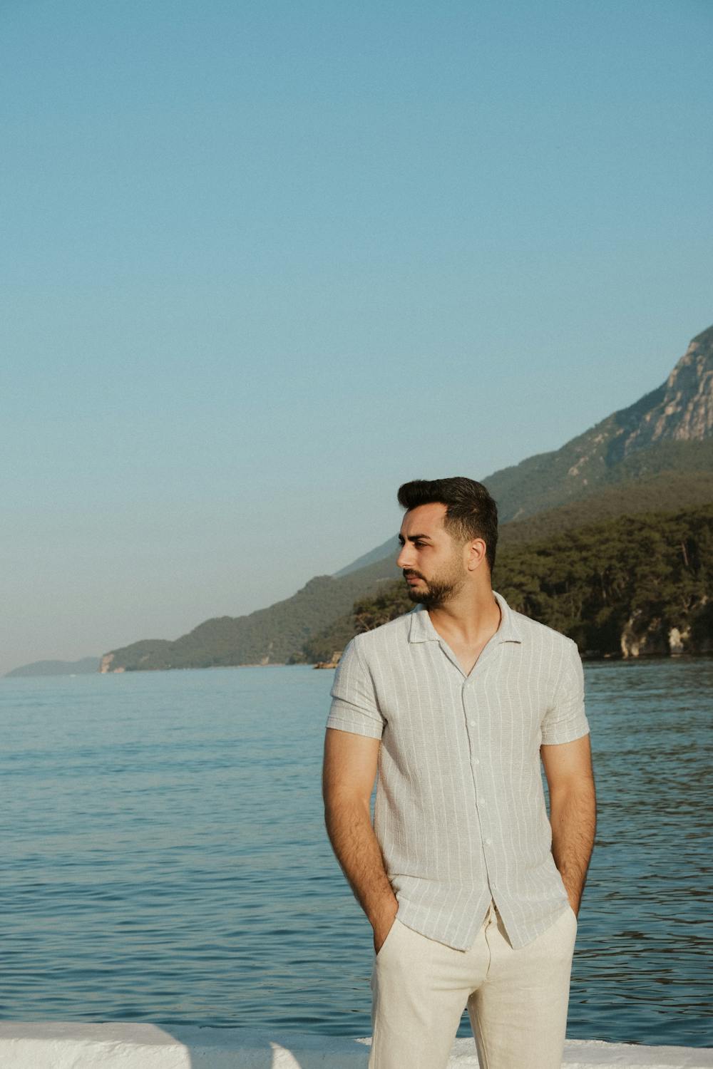 Man in White Shirt and Pants Standing at a Waterfront with a Mountain ...