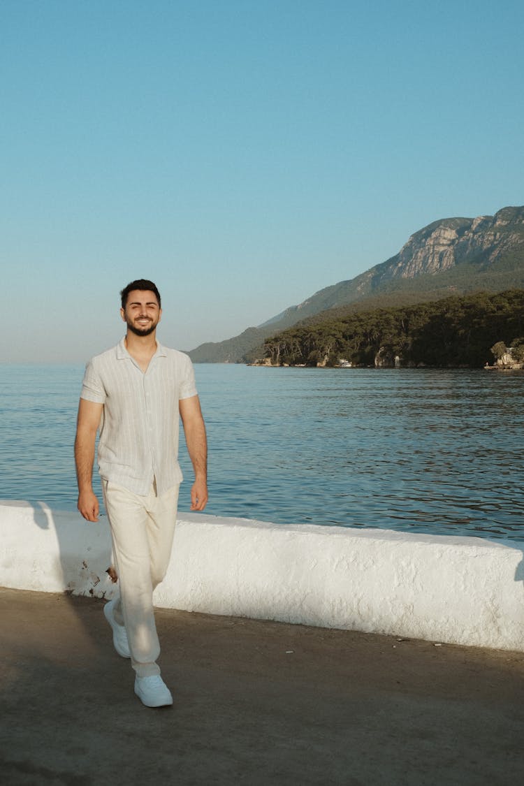 Brunette Man In White Short Sleeved Shirt And Pants Walking On A Sea Waterfront