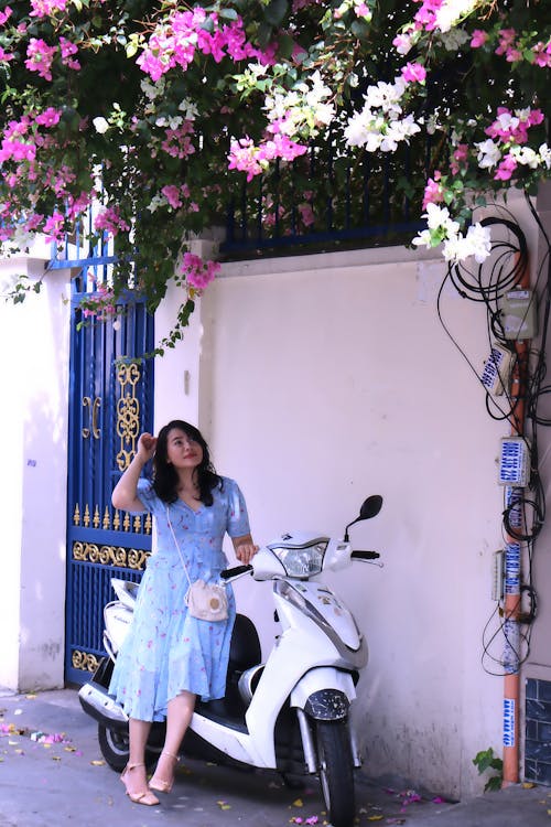 Young Woman Leaning against a Motor Scooter and Looking at Flower above the Fence 