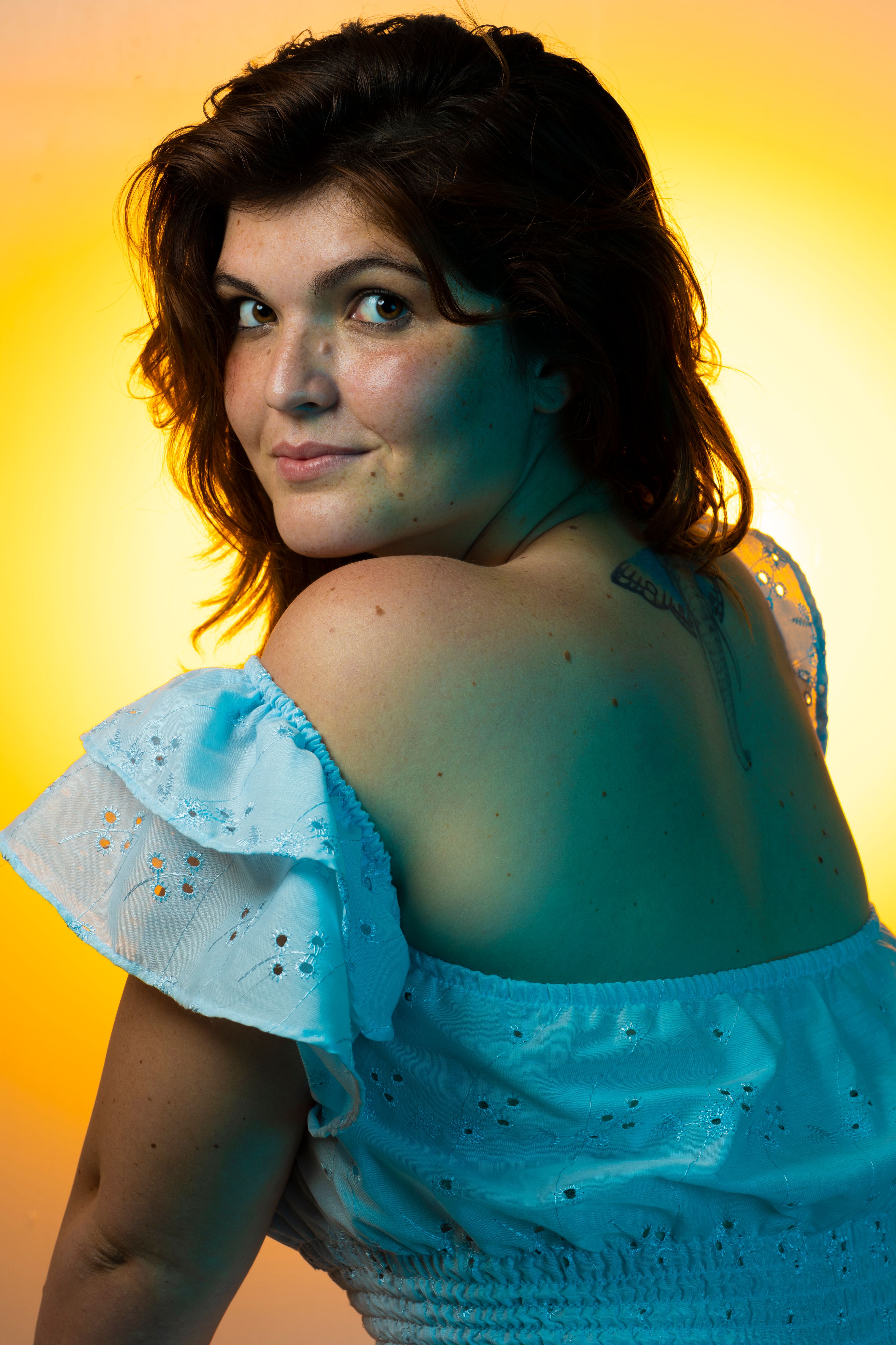 Portrait of a woman with brown hair, smiling in a white dress against a warm yellow background.