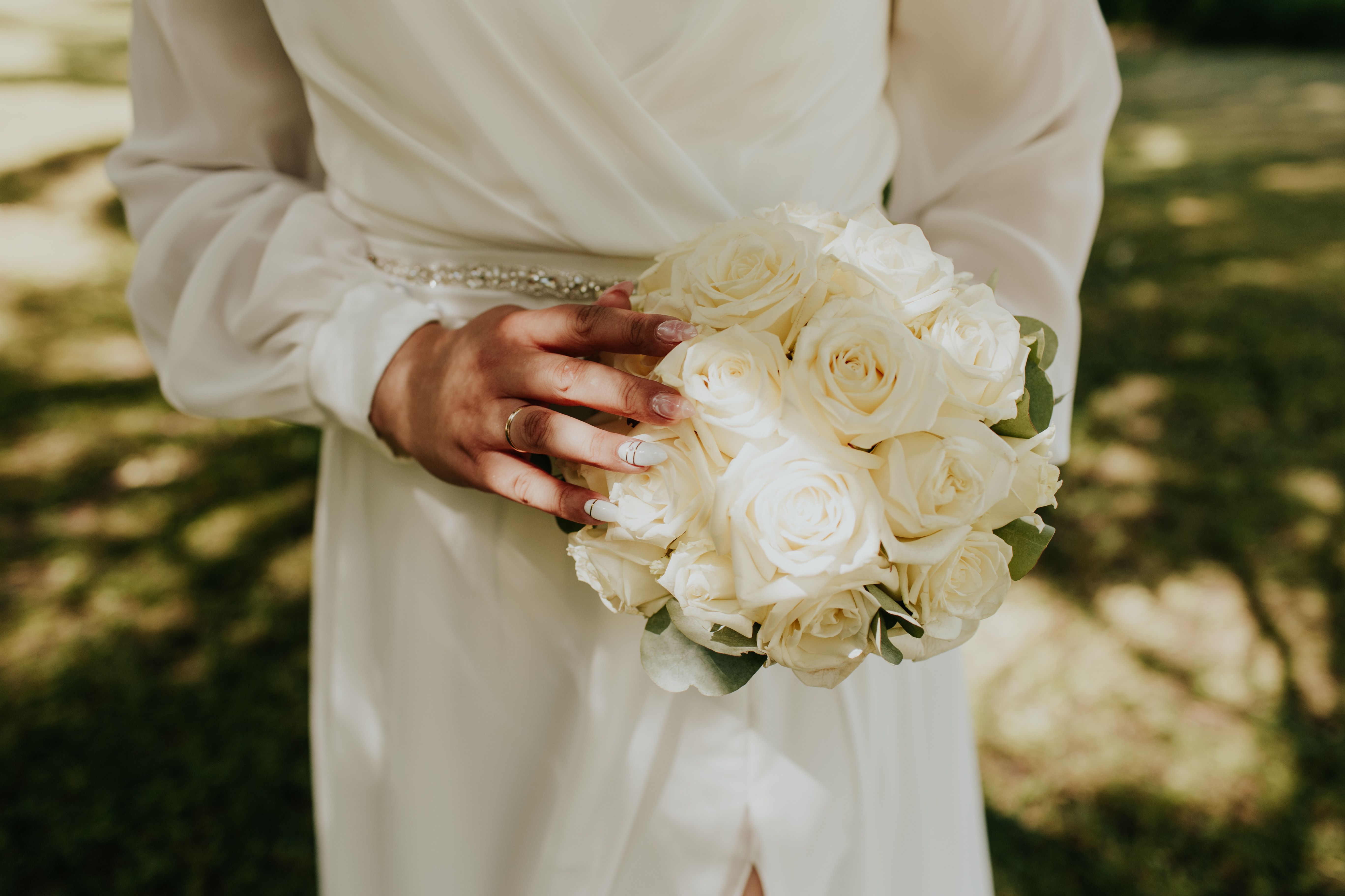 Bride holding a bouquet of white roses outdoors in sunlight, showcasing elegance and beauty.