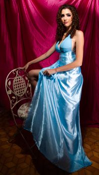 Elegant woman in a blue dress posing with a chair against a red background.