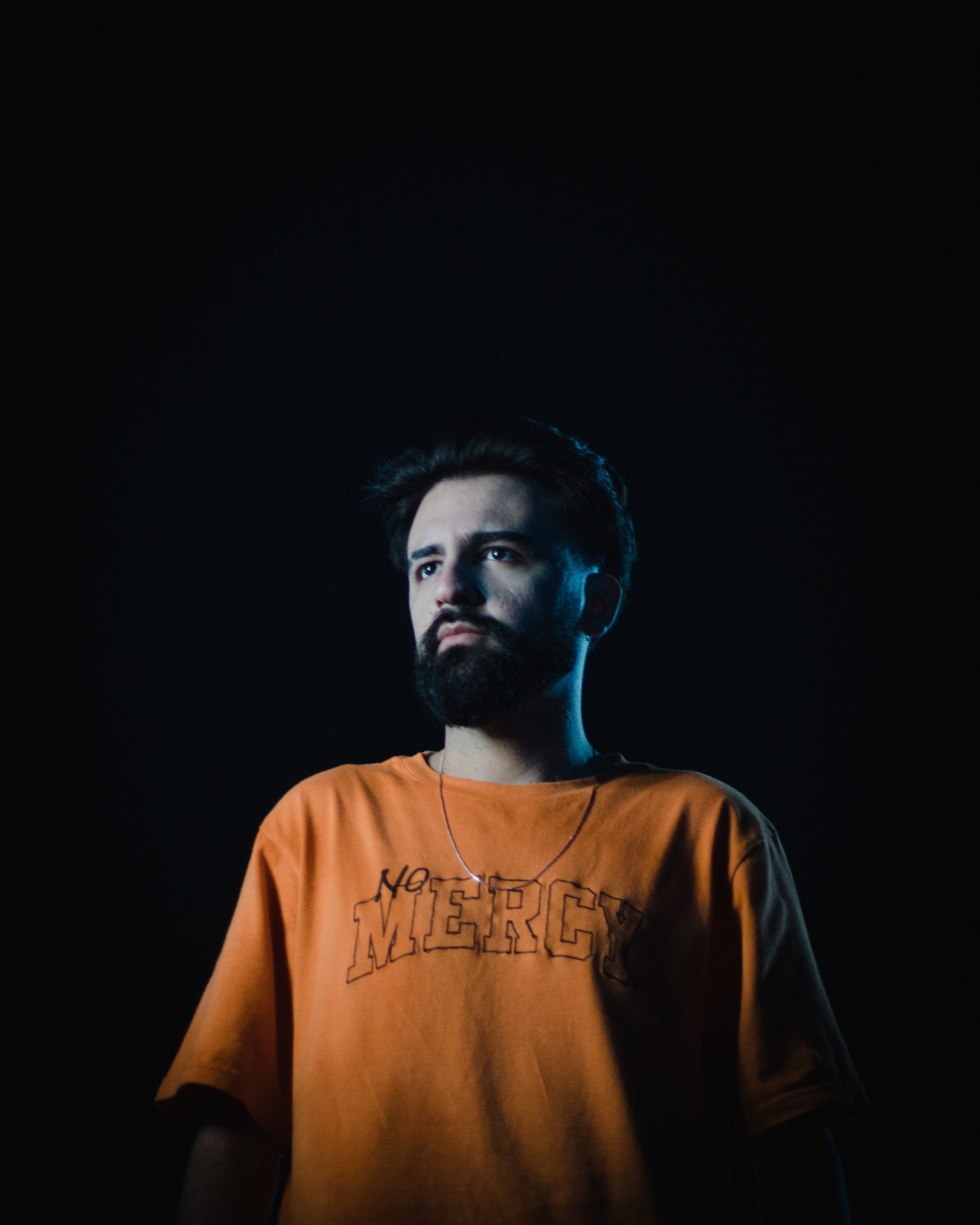 Dramatic portrait of bearded man wearing 'No Mercy' t-shirt against dark background.