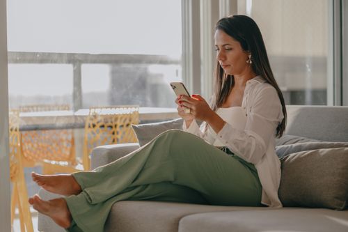 Free A woman in a white shirt and green pants uses her cellphone on a gray couch indoors. Stock Photo