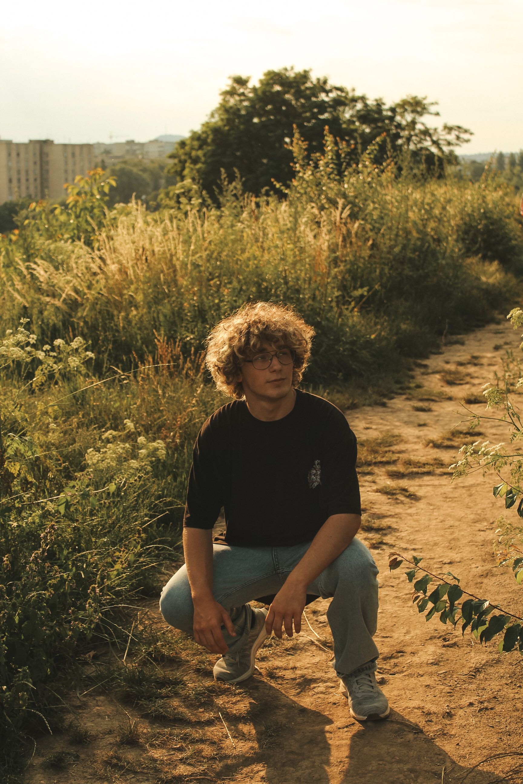An adult male poses crouching on a rural path surrounded by lush greenery in warm lighting.