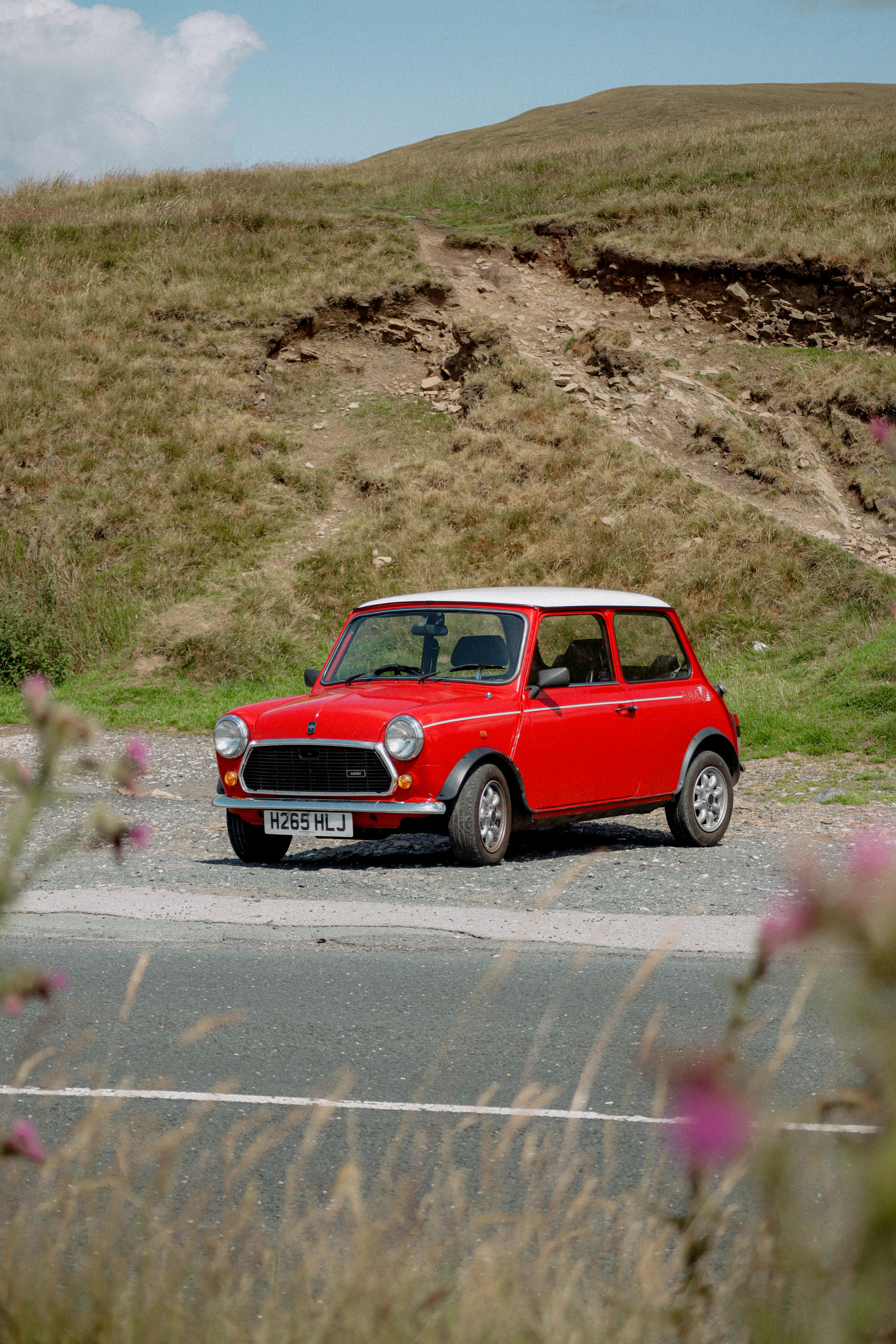 Vintage red Mini Cooper parked in a picturesque rural landscape with rolling hills in the background.