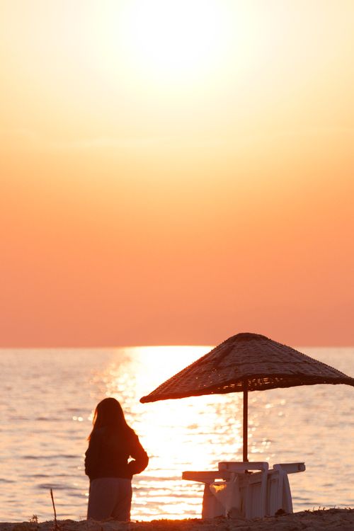 Free Silhouette of a woman at sunset on a beach with a straw umbrella, embracing tranquility. Stock Photo