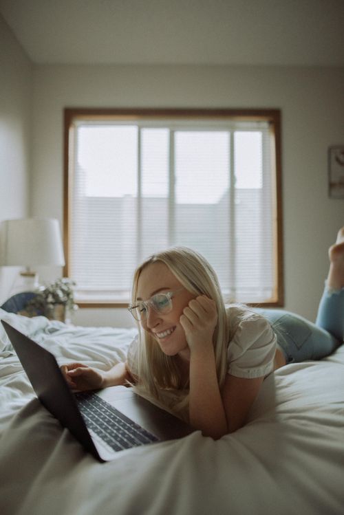 Free A happy woman with long blonde hair in eyeglasses lying on a bed using a laptop in a bright room. Stock Photo