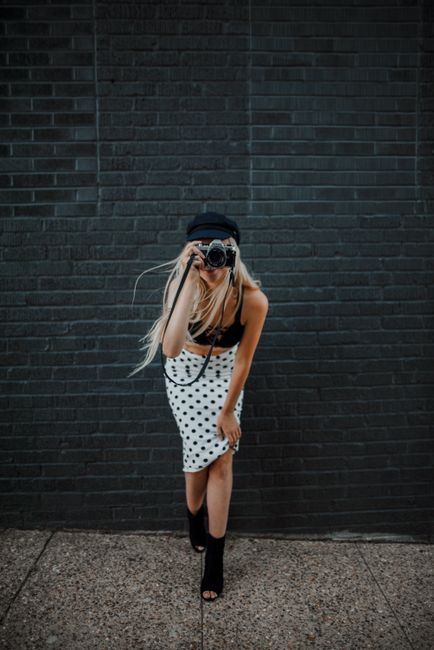 Blonde woman in polka dot skirt with camera posing outside city wall.