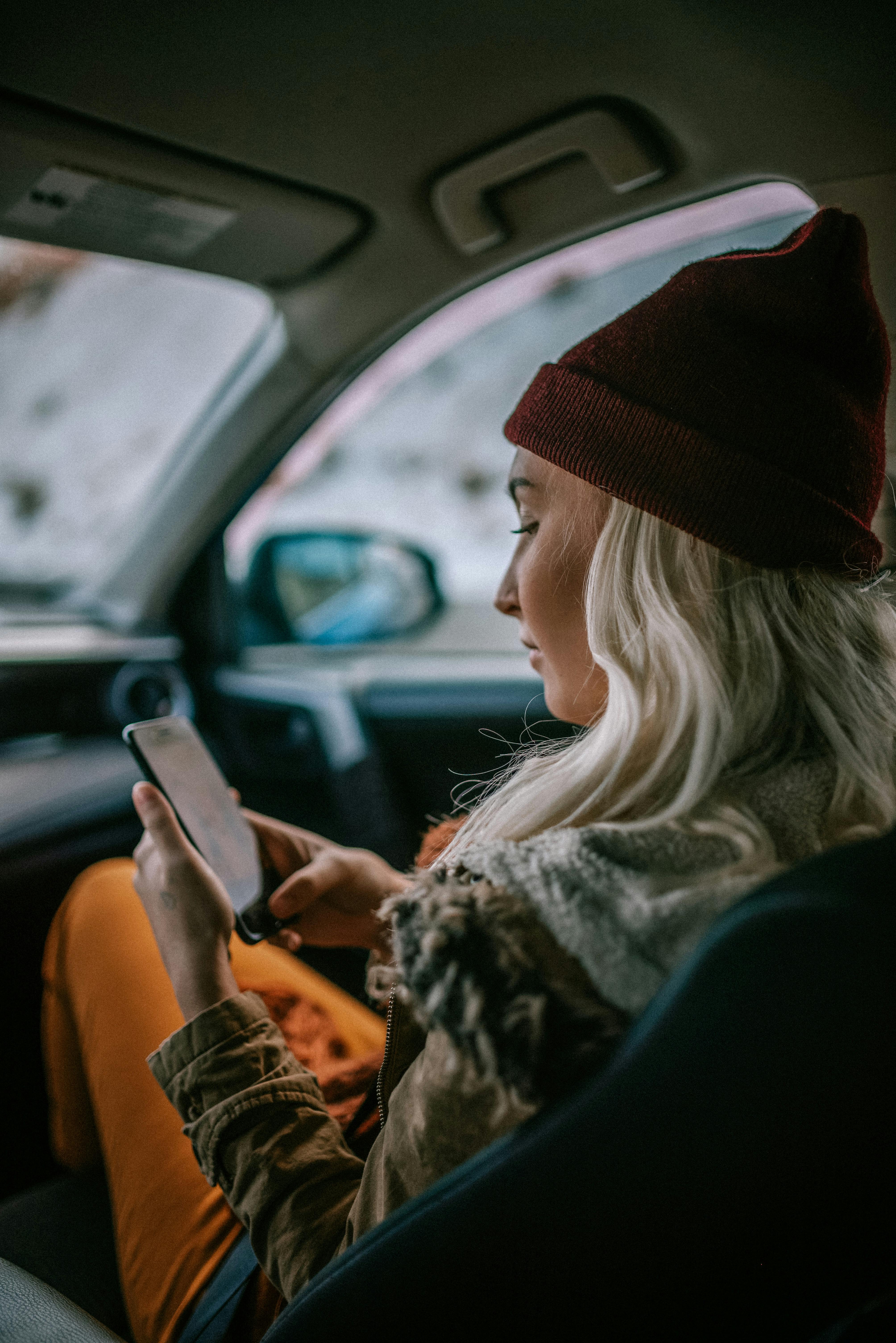 A woman with blonde hair and a beanie using a smartphone inside a car during winter travel.