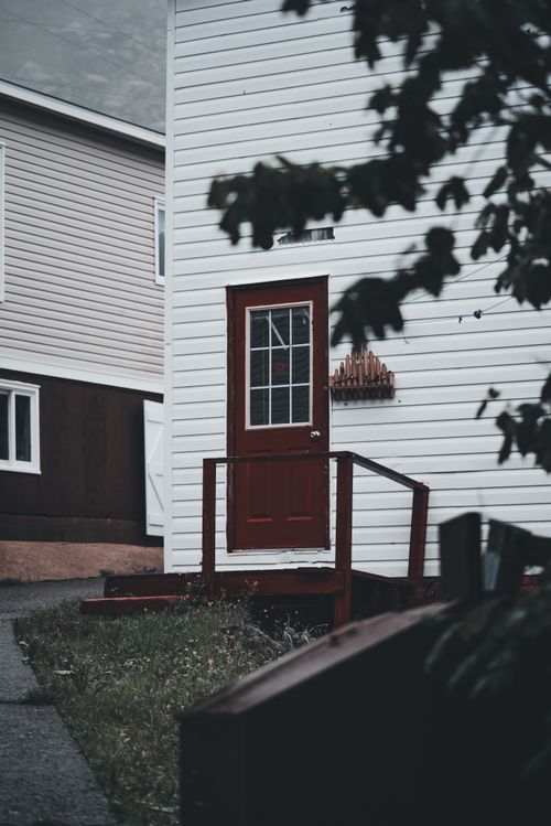 Free A quaint white house with a striking red door, nestled among greenery, captures a peaceful rural setting. Stock Photo