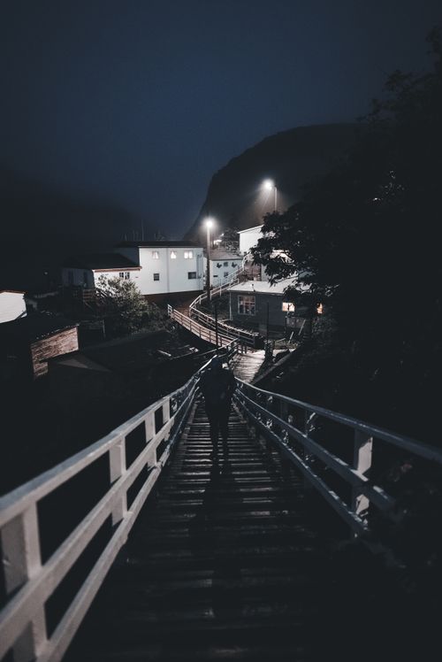Free Silhouette of a person walking along a village boardwalk at night, illuminated by streetlights. Stock Photo