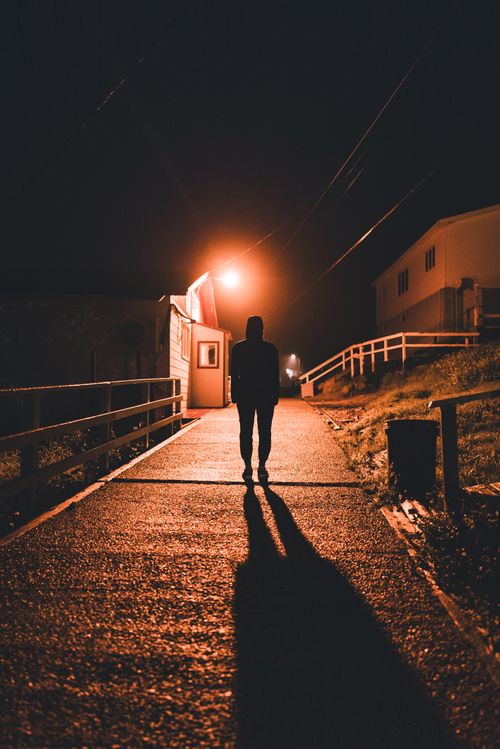 Free A lone figure stands silhouetted under a streetlight in a quiet village setting at night. Stock Photo