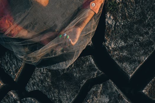 A close-up shot of a woman's painted nails under a veil on cracked stone pavement, evoking a concept of vulnerability and beauty.