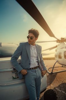 Man in a suit stands by a helicopter and car at sunset, exuding elegance.