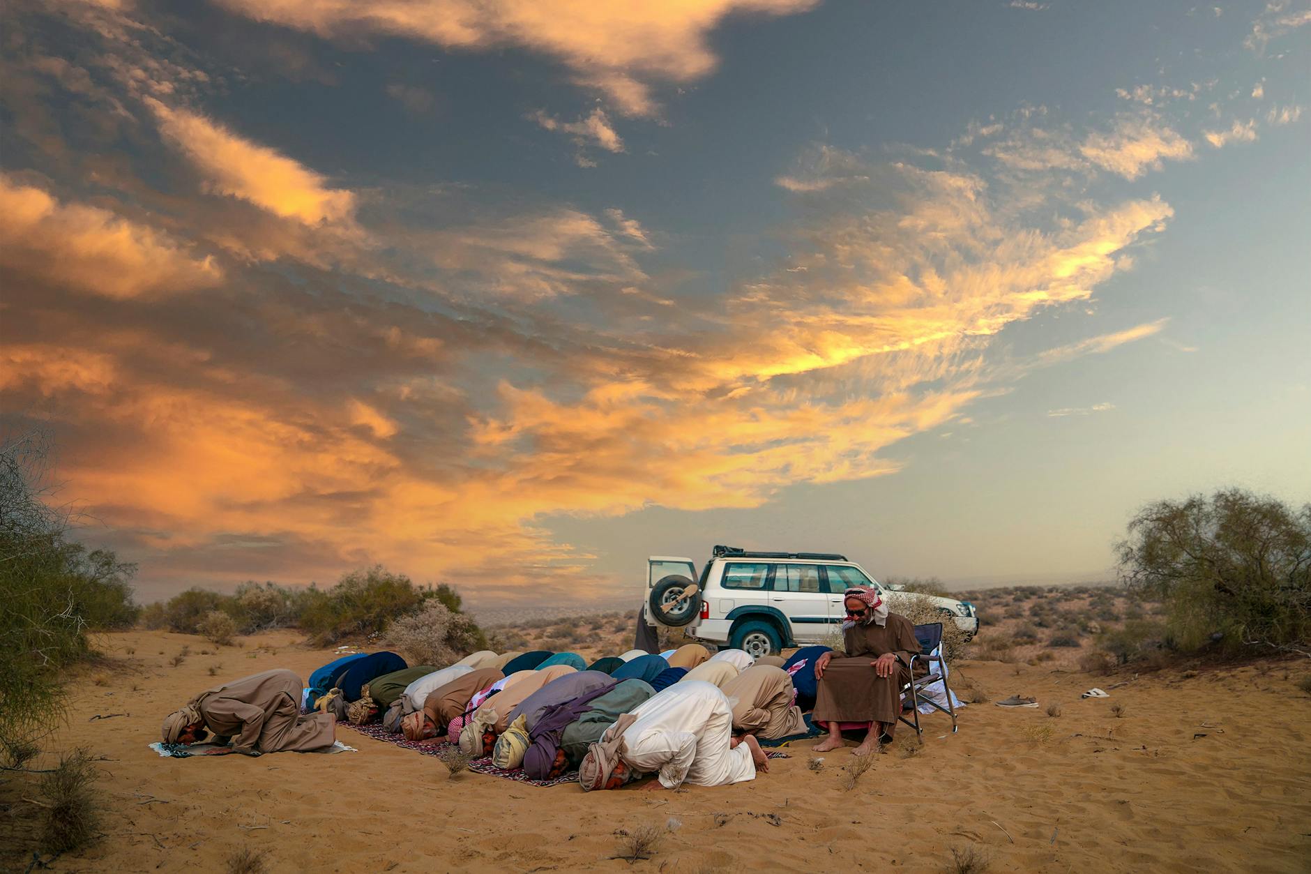 Group of men in traditional attire praying in the desert at sunset.