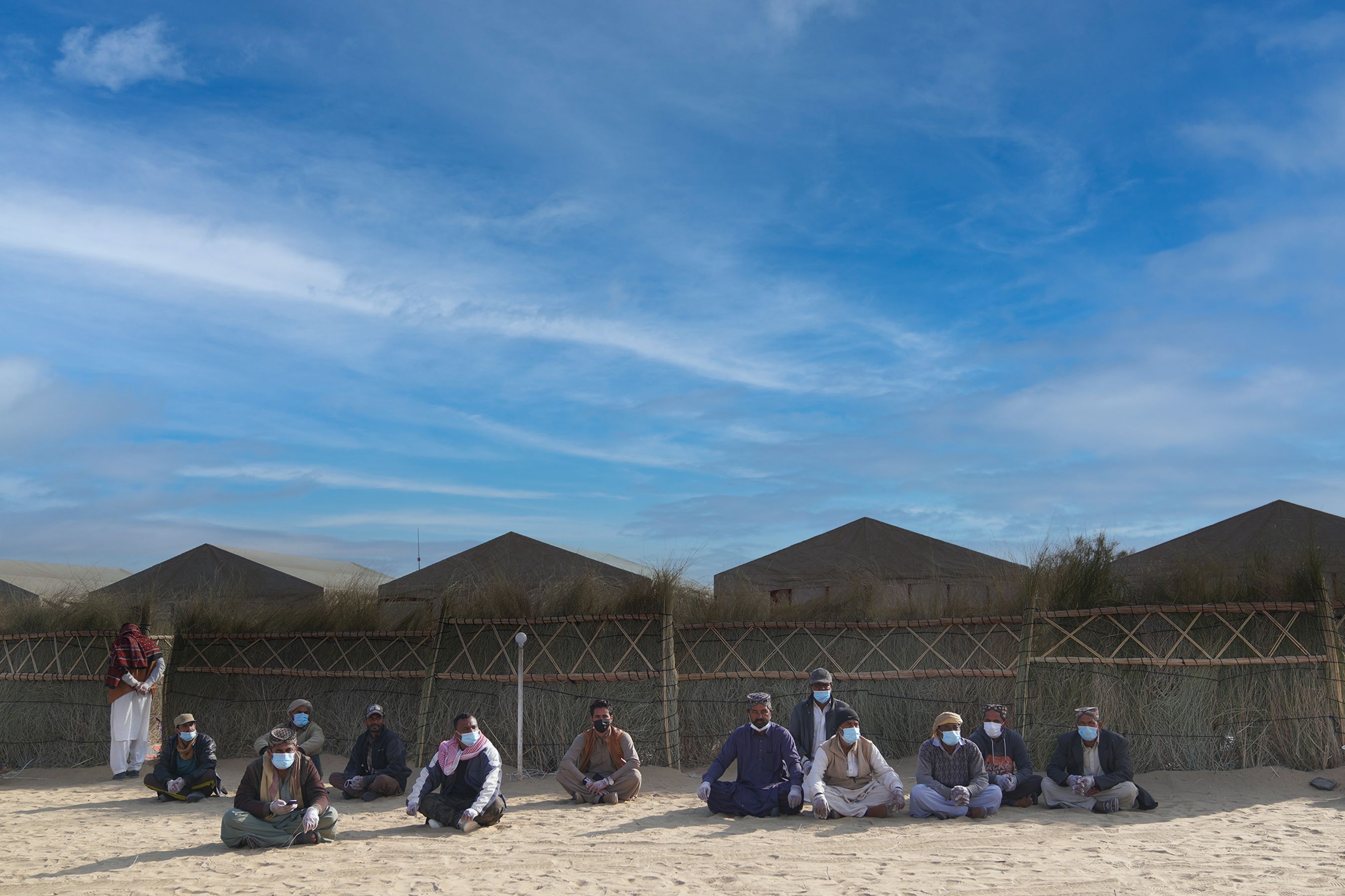 A group of men sitting on sand in a desert camp setting under a blue sky.