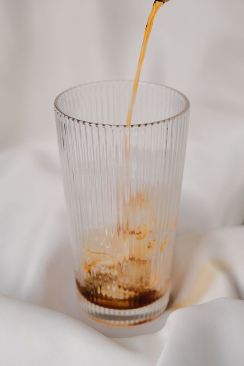 Free A vertical shot of coffee being poured into a tall, clear glass set on a white cloth, creating a minimalist aesthetic. Stock Photo