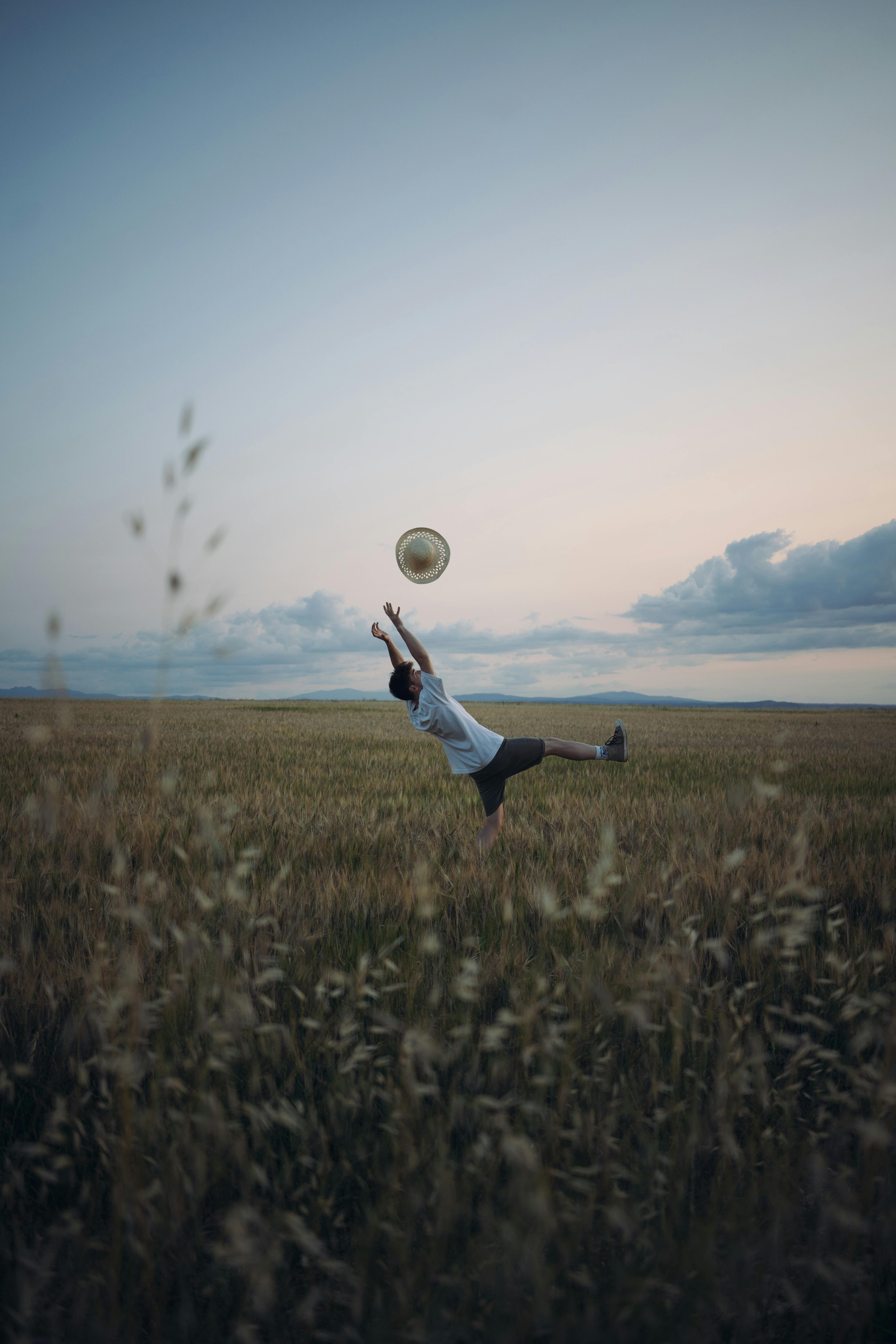 Young boy throws a summer hat in a grassy field during twilight, showcasing playful joy.