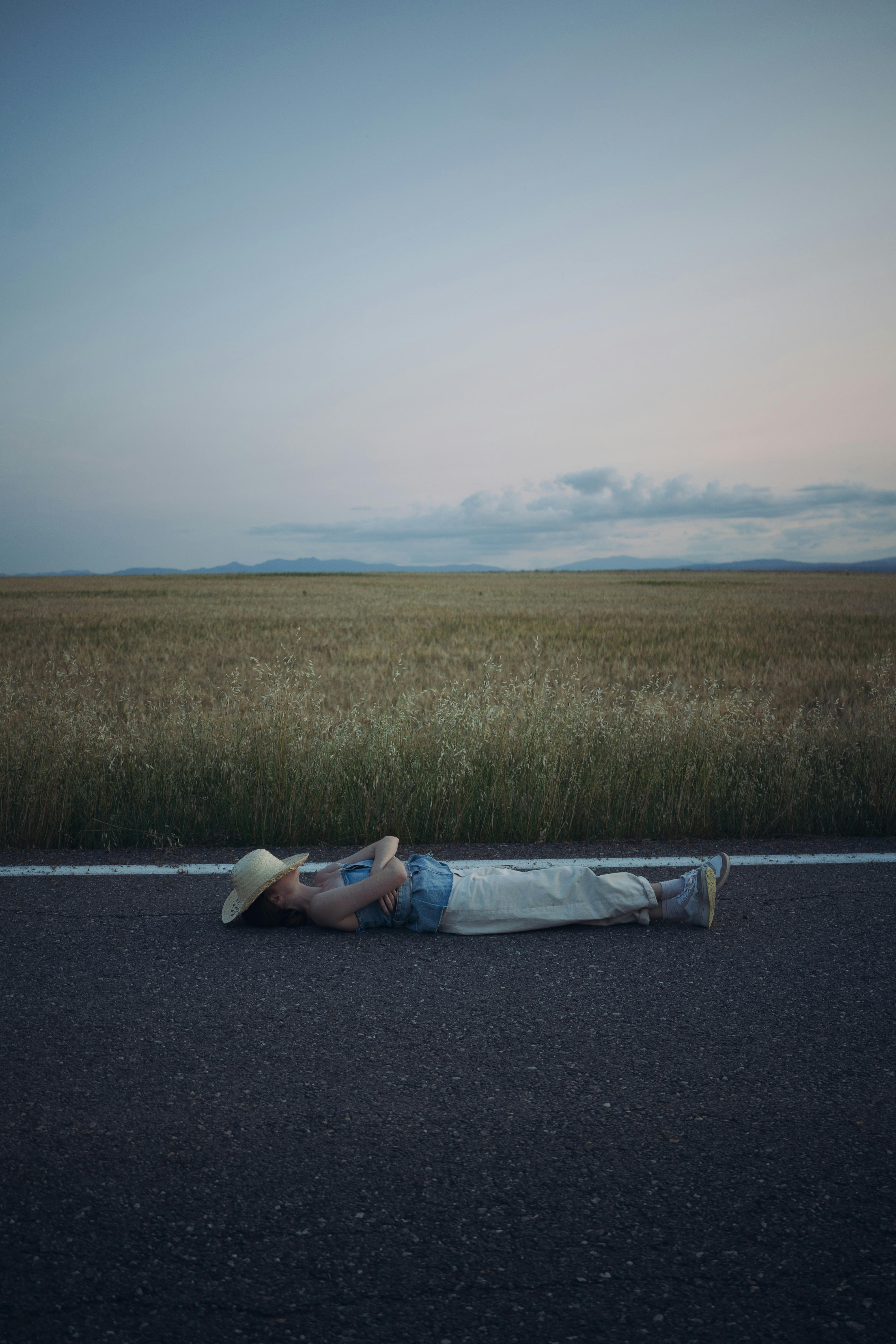A woman in casual clothes and a hat lays peacefully on a country road at twilight.