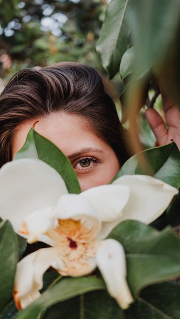 A woman with brunette hair peeks through lush green leaves and a large white magnolia flower.
