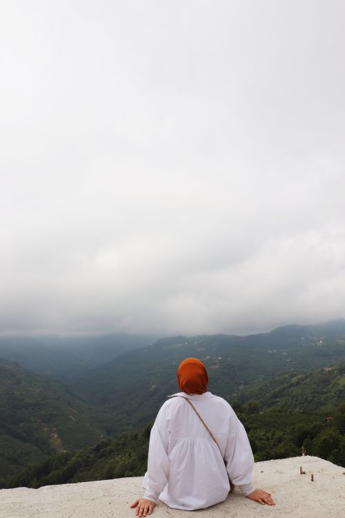 Free A woman in a hijab sitting on a ledge, gazing over a cloudy, lush valley, exuding serenity and reflection. Stock Photo