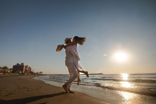 Free A couple in white attire joyfully embracing on a sunlit beach with a distant city view. Stock Photo