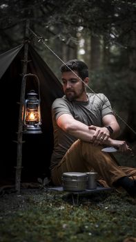 Man camping in Bolu forest, sitting by tent with vintage lantern and camping gear.