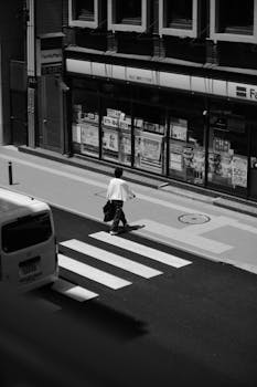 Black and white high-angle view of a person crossing a city street crosswalk near a storefront.