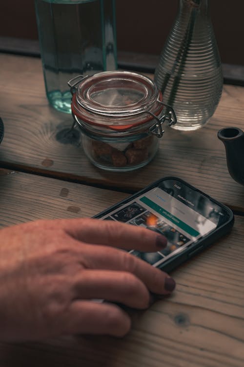 Hand of a Woman Holding a Smartphone and a Jar on a Table 