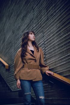 Fashionable woman with wavy hair standing on stairs in stylish outfit, urban background.