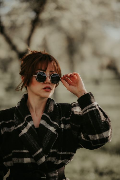 Free Fashionable young woman in a checked coat posing with sunglasses in a serene park setting. Stock Photo