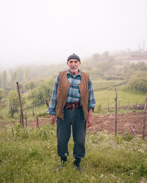 Free An elderly farmer in a checkered shirt stands in a foggy rural field, surrounded by hills and grass. Stock Photo