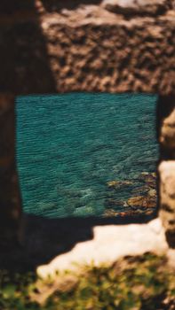 A beautiful view of the clear sea framed by a rustic stone window, captured in summer.