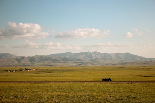 A car drives along a road through open fields with mountains in the background, under a clear sky.
