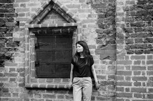 Woman Leaning Against the Wall of an Old Brick Building Next to a Shuttered Window