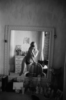 Black and white photo of a woman capturing her reflection in a mirror inside a room.
