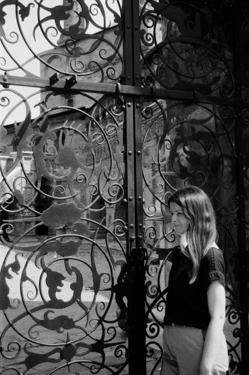 Young Woman in Black T-shirt Posing by Ornately Decorated Iron Gates