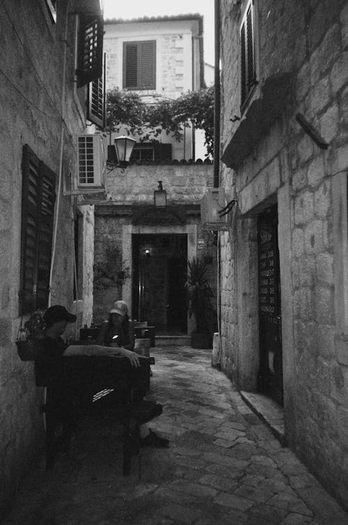 Black and White Photo of People Sitting by Outdoor Cafe Tables in Narrow Street
