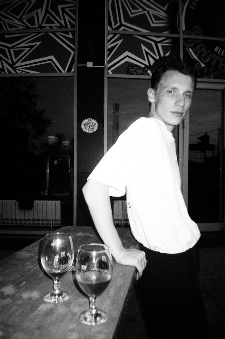 Black And White Photo Of A Young Man Leaning On A Bar Stand With Glasses