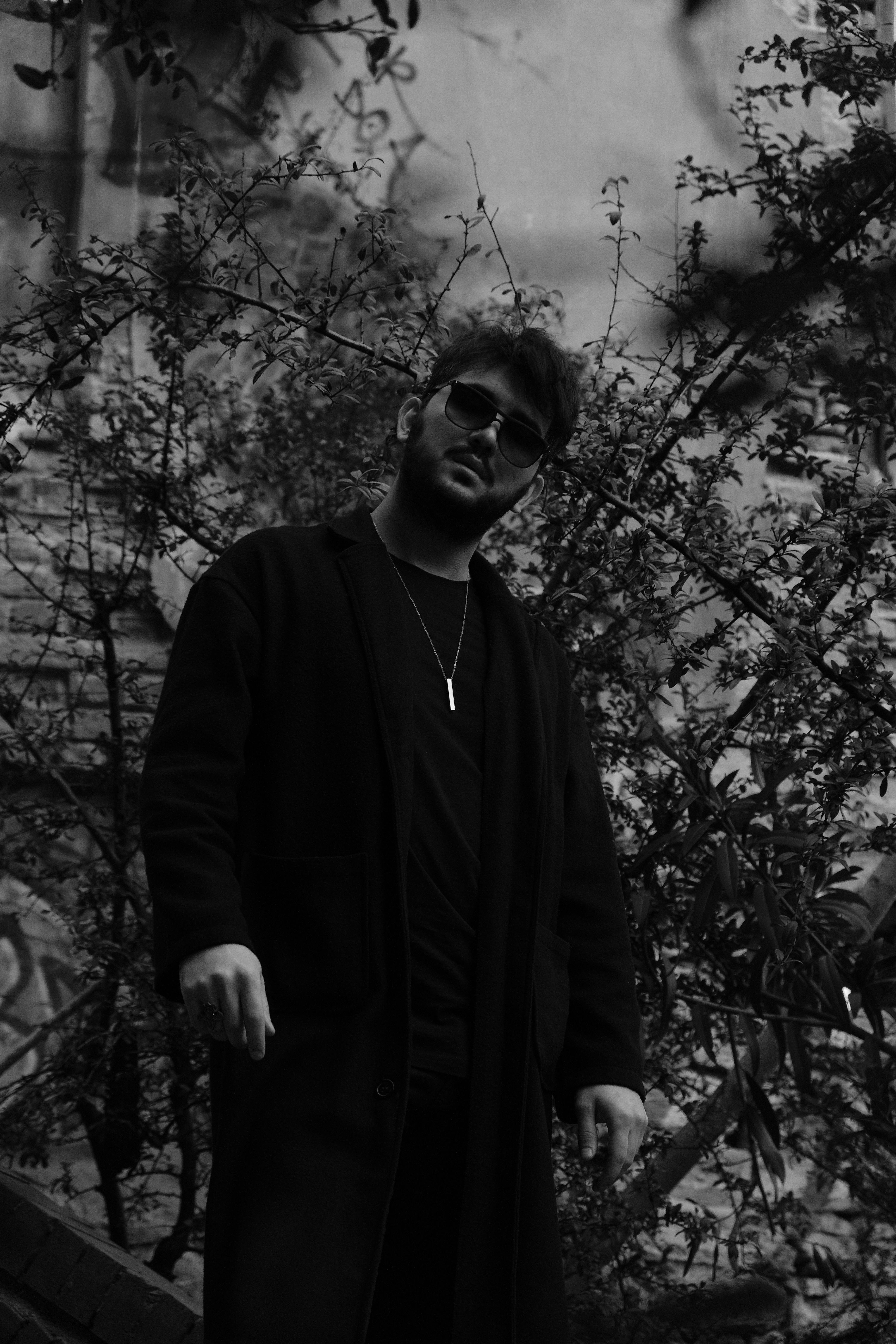 Urban portrait of a man in black attire with sunglasses, surrounded by foliage.