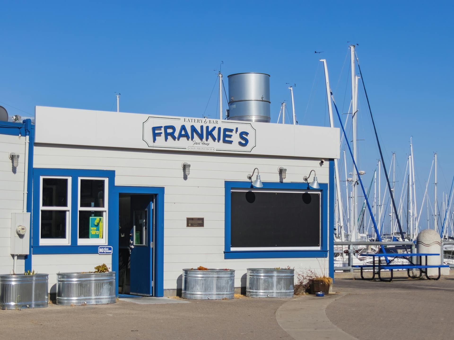Sunlit seaside eatery and bar located by a marina with sailboats docked nearby.
