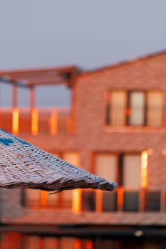 Close-up of a wicker umbrella with sunset reflections on a modern building facade.