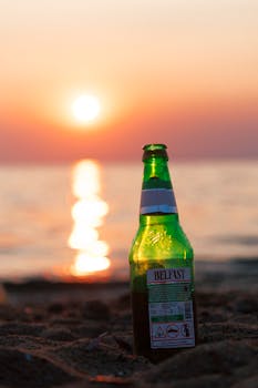 Green beer bottle on sandy beach with ocean sunset backdrop, ideal for summer vibes.