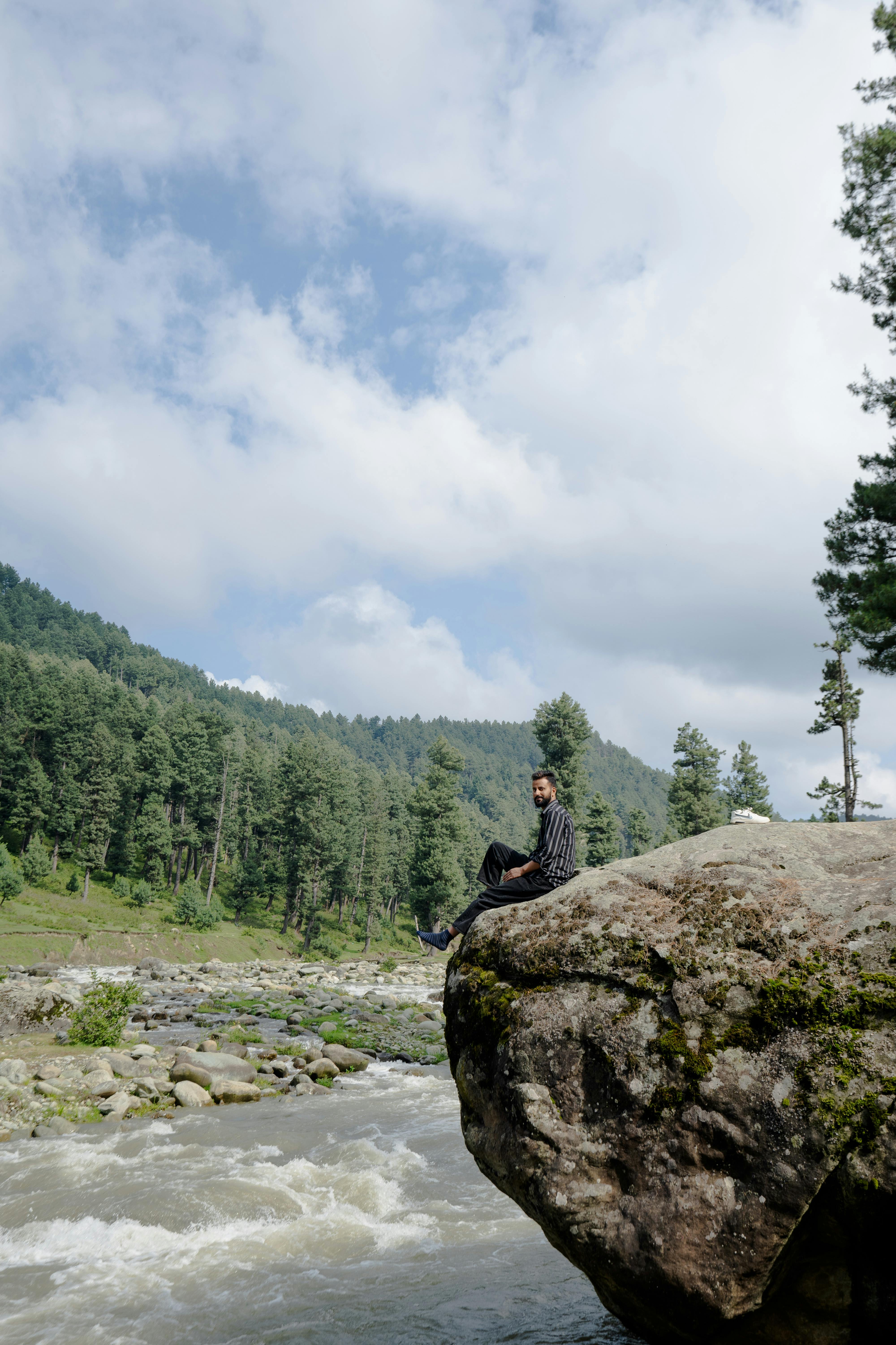 A man sitting on a large rock overlooking a flowing stream in a forested area, capturing serenity.