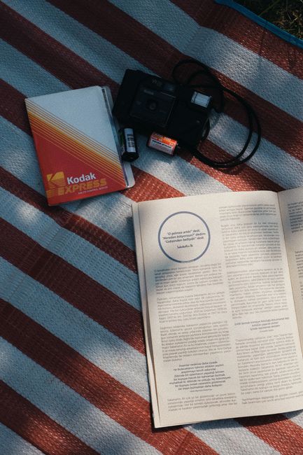 A nostalgic outdoor setup with a Kodak camera, film, and a book on a striped blanket in daylight.