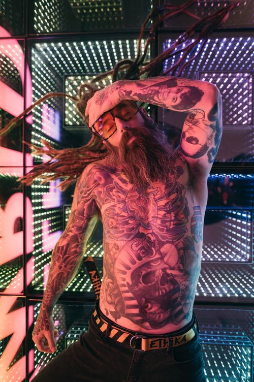 Free Energetic shot of a shirtless, tattooed man posing with vibrant neon lights and dreadlocks. Stock Photo