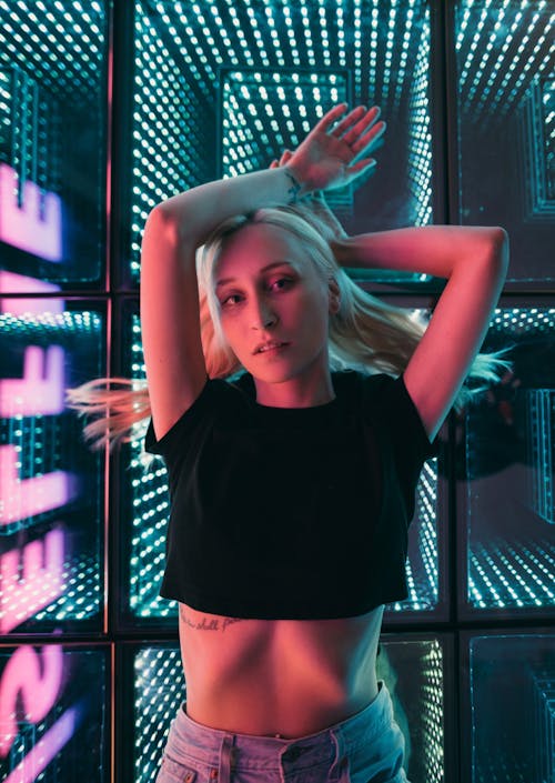 Young Blonde Woman in Black Crop Top Lying on a Glass Floor Lit by LED Lamps